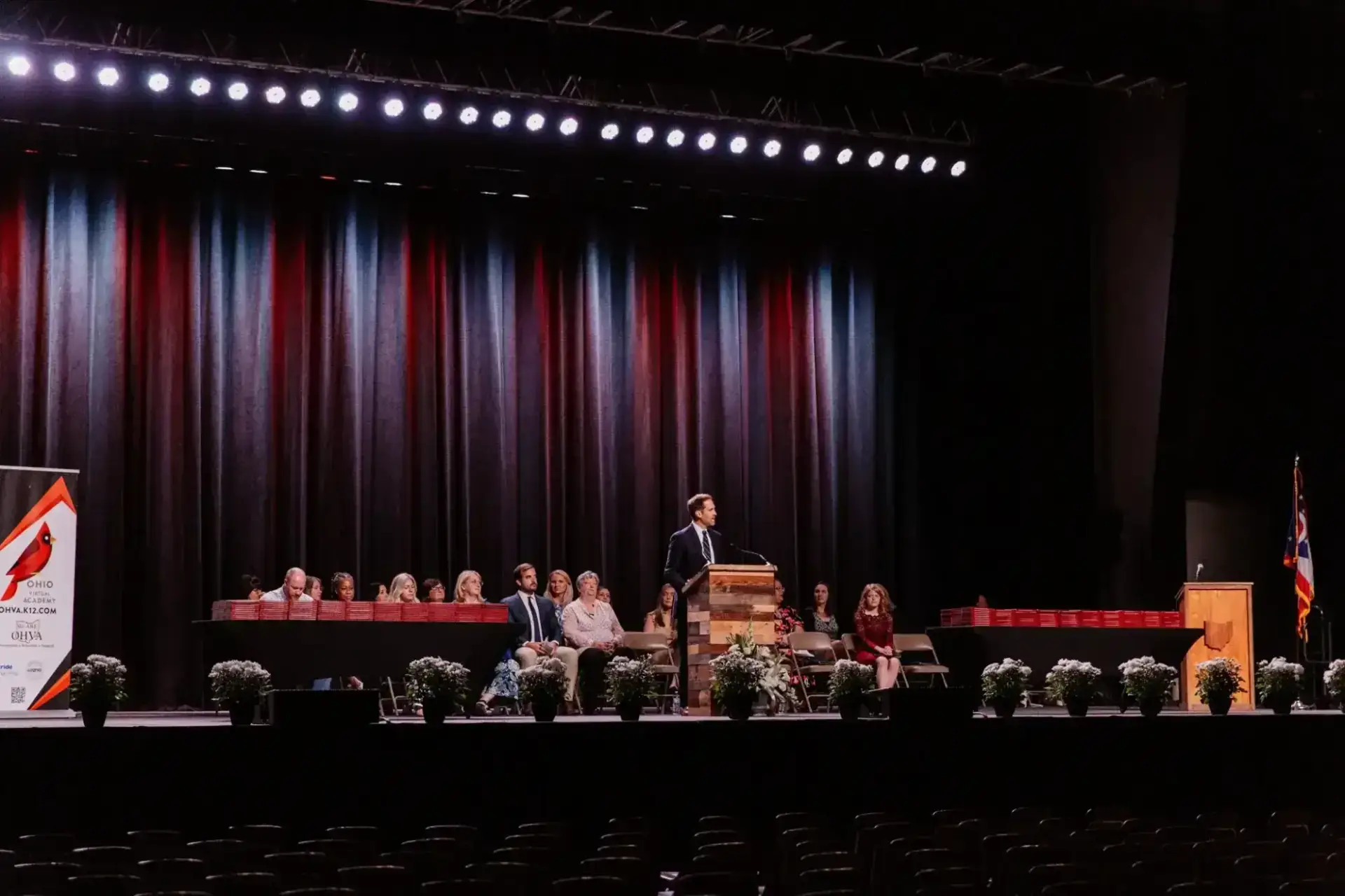 رجل يقف على منصة على مسرح كبير مع ستارة خلفية، يخاطب الجمهور. يصطف على المسرح أشخاص جالسون وأصص نباتات. الأجواء رسمية.