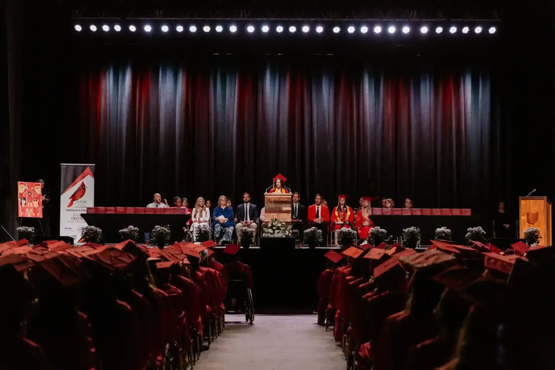 حفل التخرج حيث يجلس الطلاب مرتدين القبعات والعباءات الحمراء في مواجهة المنصة. متحدث على المنصة يحيط به أعضاء هيئة التدريس.