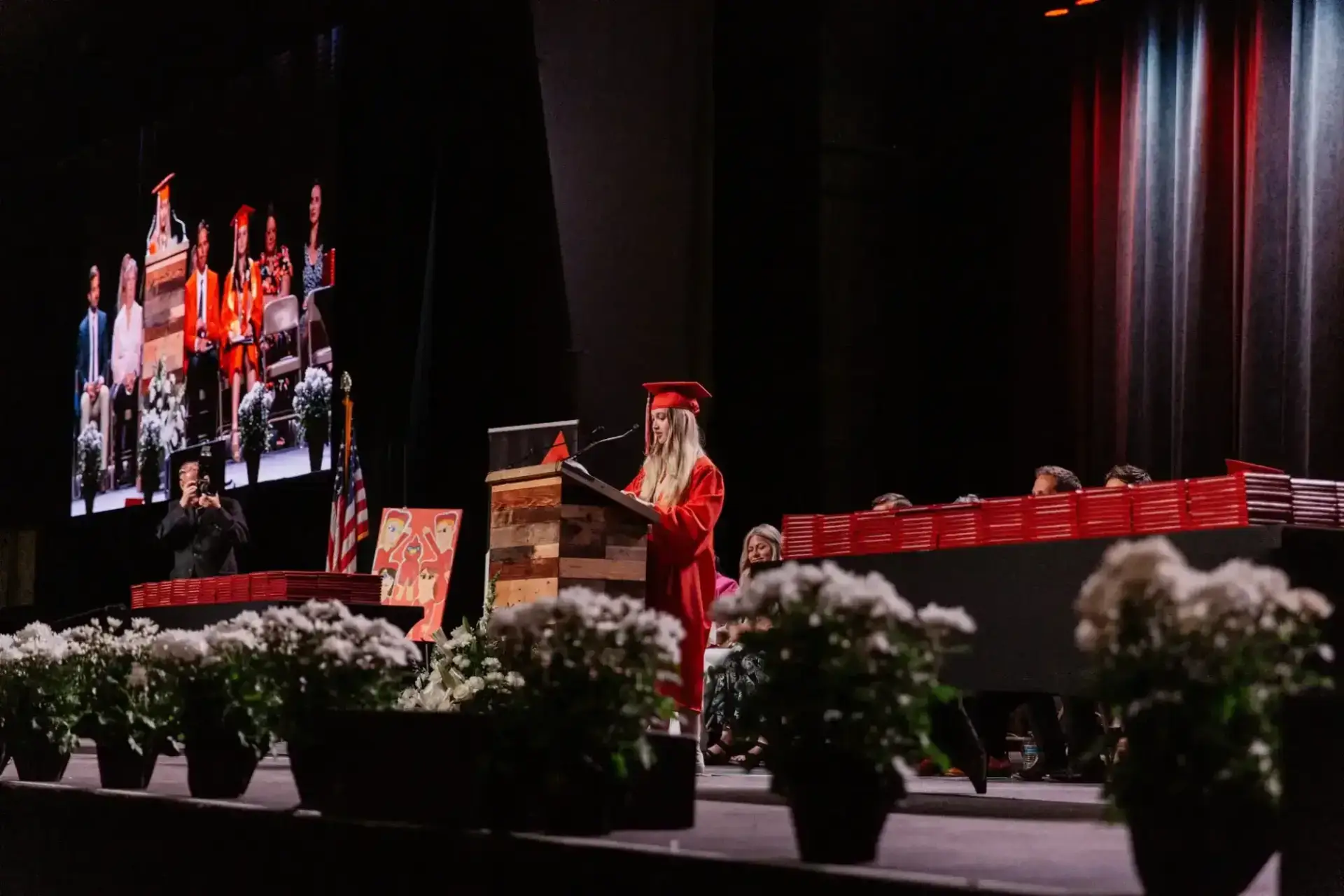 طالب يرتدي قبعة حمراء وعباءة حمراء يتحدث على منصة خشبية على مسرح التخرج المضاء إضاءة خافتة، مع ترتيبات زهرية وخلفية شاشة كبيرة.