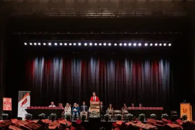 يقف متحدث يرتدي رداءً أحمر على منصة على خشبة المسرح، ويراقبه جمهور من الخريجين الذين يرتدون قبعات حمراء.