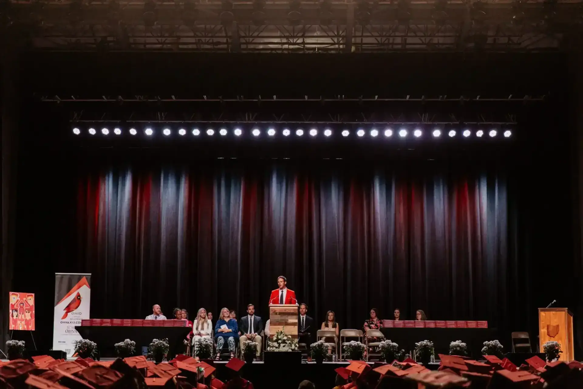 يقف متحدث يرتدي رداءً أحمر على منصة على خشبة المسرح، ويراقبه جمهور من الخريجين الذين يرتدون قبعات حمراء.