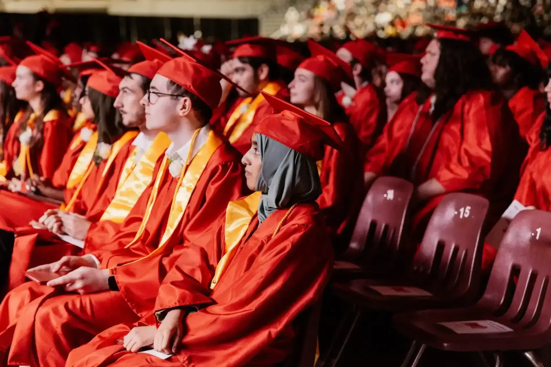 خريجون يرتدون قبعات حمراء وعباءات حمراء مع شالات صفراء يجلسون في صفوف خلال الحفل.