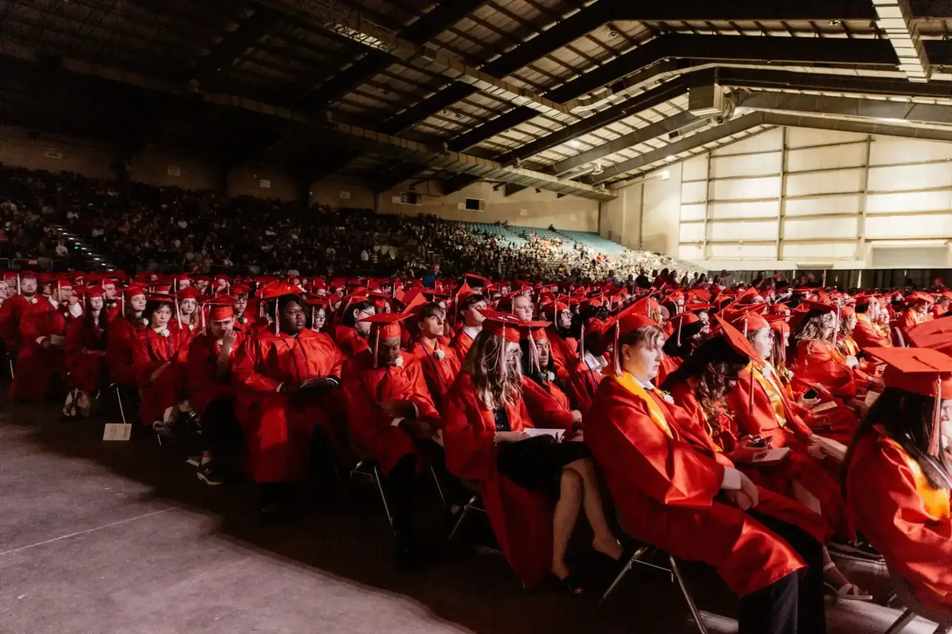 مجموعة كبيرة من الطلاب الذين يرتدون القبعات والعباءات الحمراء يجلسون في صفوف خلال حفل التخرج داخل قاعة فسيحة مليئة بالمتفرجين.