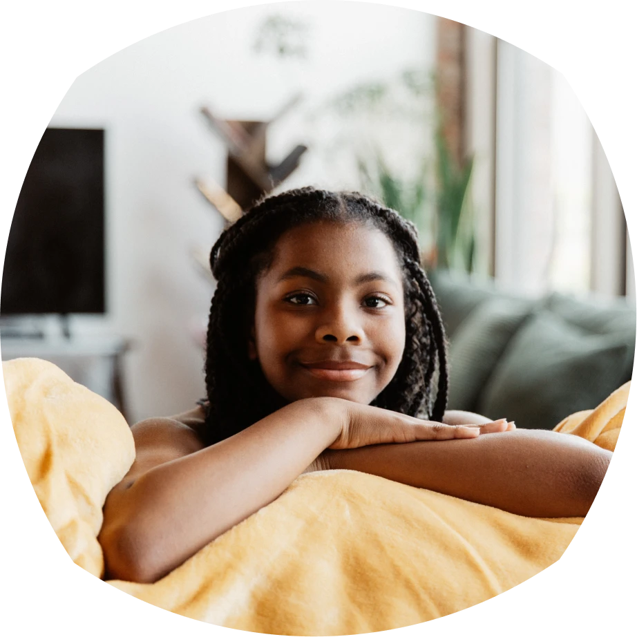 فتاة صغيرة بشعر مجدول تتكئ على أريكة وتبتسم بحرارة. وهي ملفوفة ببطانية صفراء، وفي الخلفية غرفة معيشة مريحة.