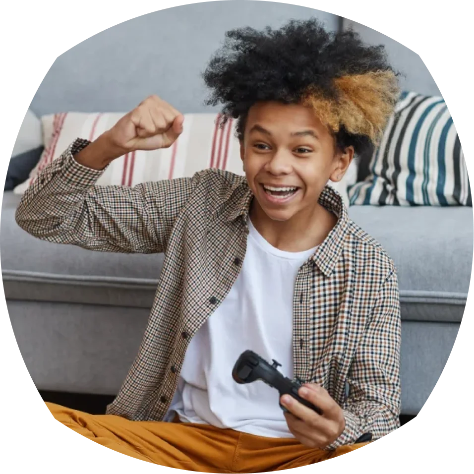 شخص شاب يرتدي قميصًا مقلّمًا وشعرًا مجعدًا يحمل بحماس وحدة تحكم في الألعاب، رافعًا قبضته وهو جالس على أريكة ذات وسائد مخططة.