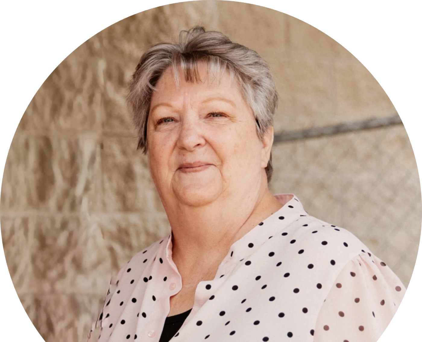 امرأة مسنة ذات شعر رمادي قصير تبتسم بهدوء وهي ترتدي بلوزة بيضاء منقطة. تُظهر الخلفية حائطاً منقوشاً باللون البيج مع سياج من السلاسل.