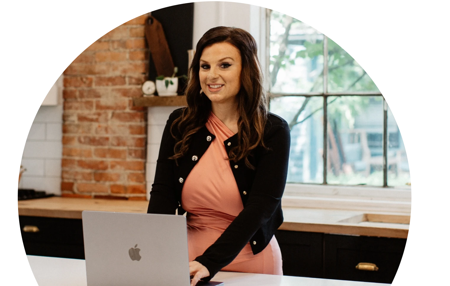 امرأة ترتدي فستانًا ورديًا وسترة سوداء تقف على طاولة المطبخ ومعها كمبيوتر محمول وتبتسم بثقة. في الخلفية جدران من الطوب ونافذة كبيرة.