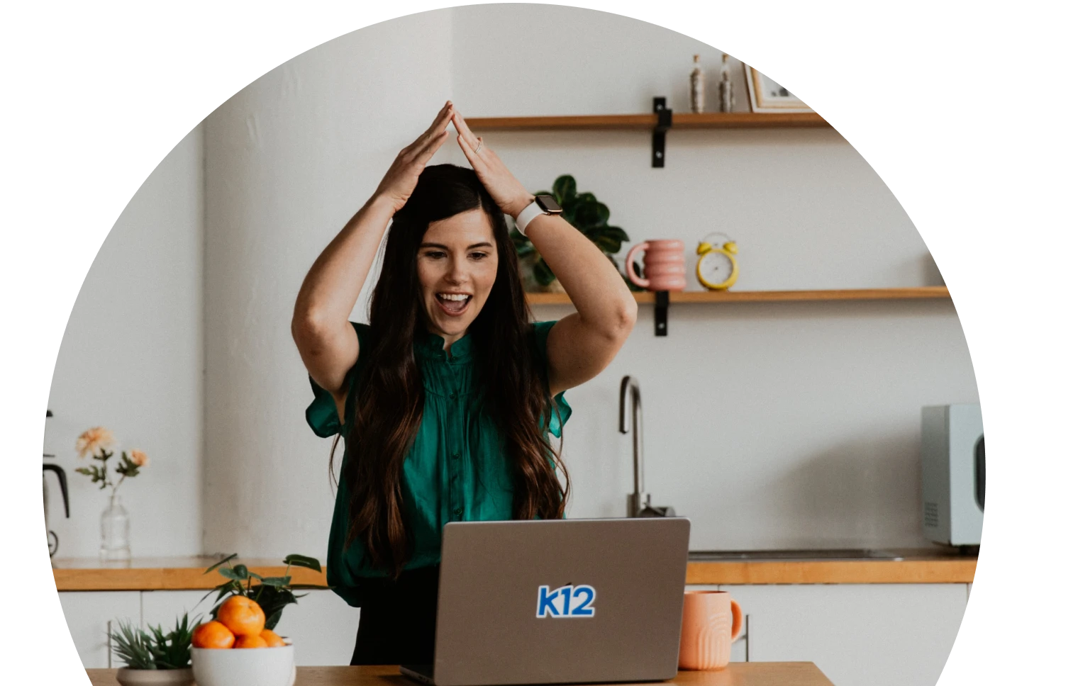 امرأة ترتدي بلوزة زرقاء مخضرة تومئ بسعادة وحماس أمام كمبيوتر محمول. الخلفية عبارة عن مطبخ مريح مع أرفف ونباتات وديكور نابض بالحياة.